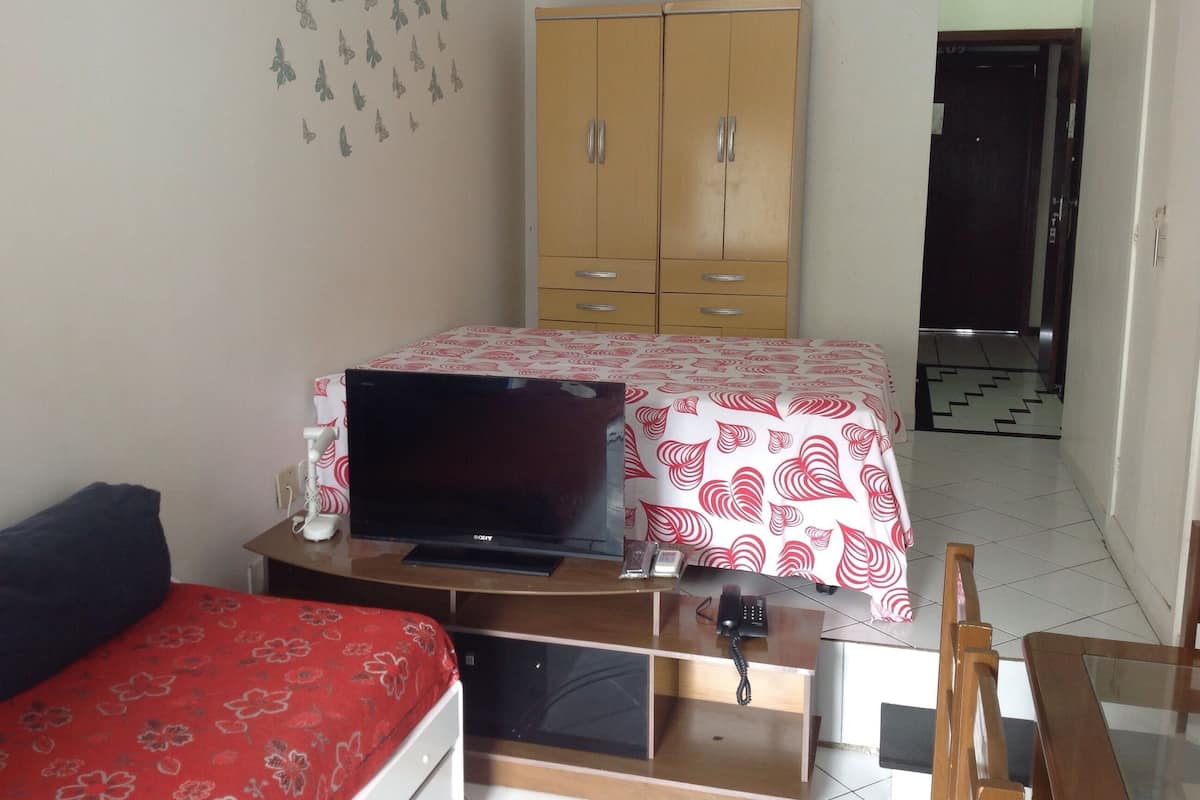 A bedroom with a bed, a desk with a laptop, a wardrobe, and a door leading to another room.