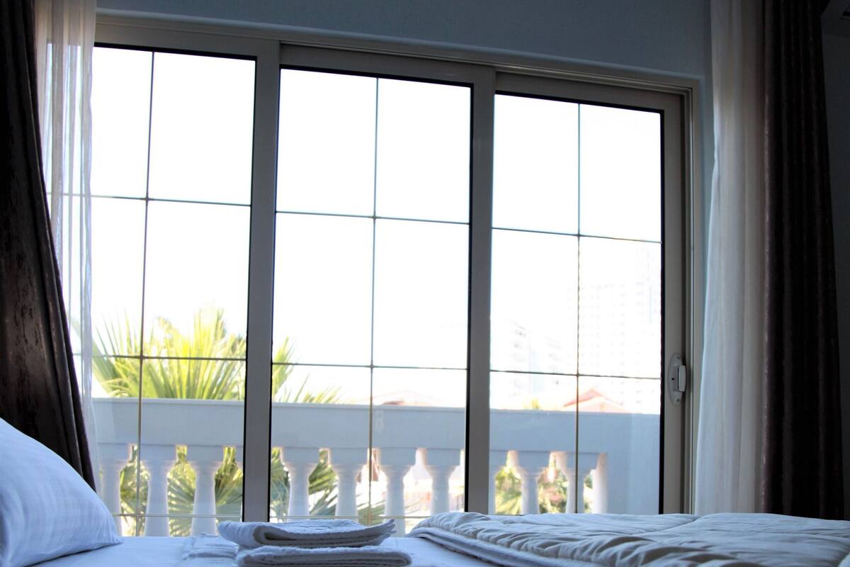 A hotel room with a large window, a bed with neatly folded white towels, and a view of palm trees and buildings outside.