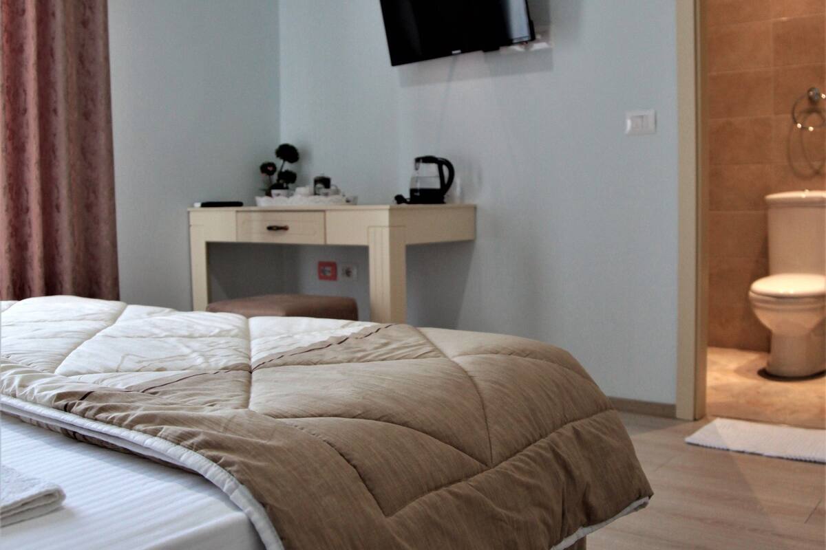 A bedroom with a bed, a desk with a flat-screen TV, an air conditioner, and a bathroom with a toilet and shower.