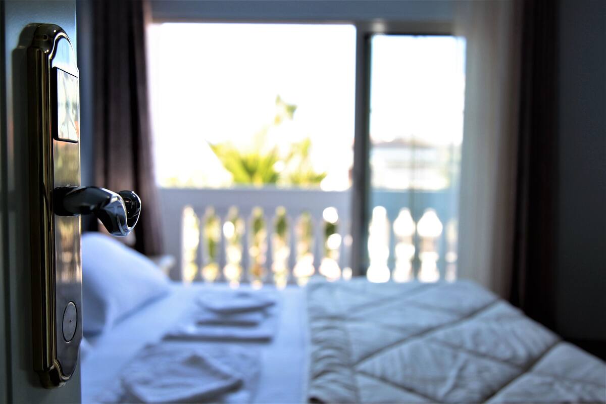 A hotel room with a bed, a window with a view of greenery, and a door with a handle.