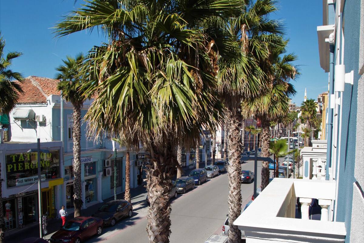 A street lined with palm trees and parked cars, buildings with various shops, and a clear blue sky.
