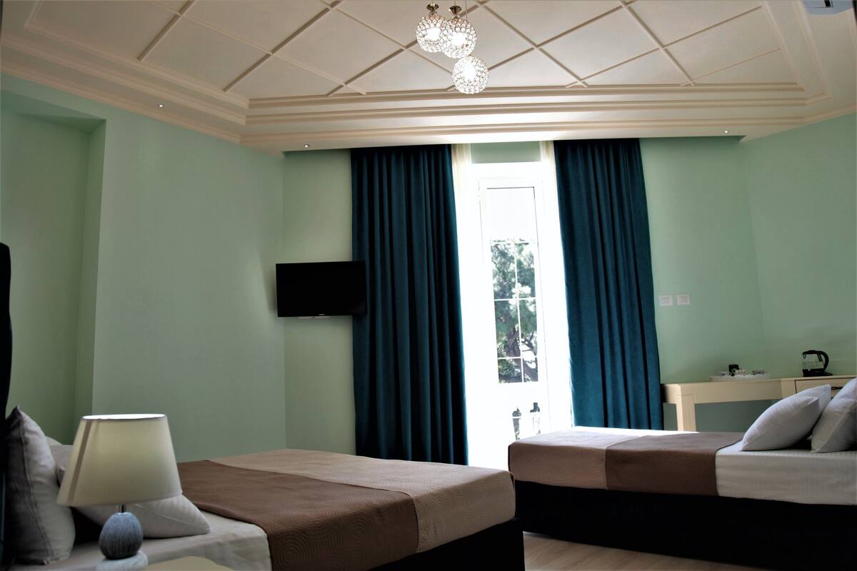A hotel room with two beds, a flat-screen TV, a desk with a lamp, and a view of the outdoors through a window with curtains.