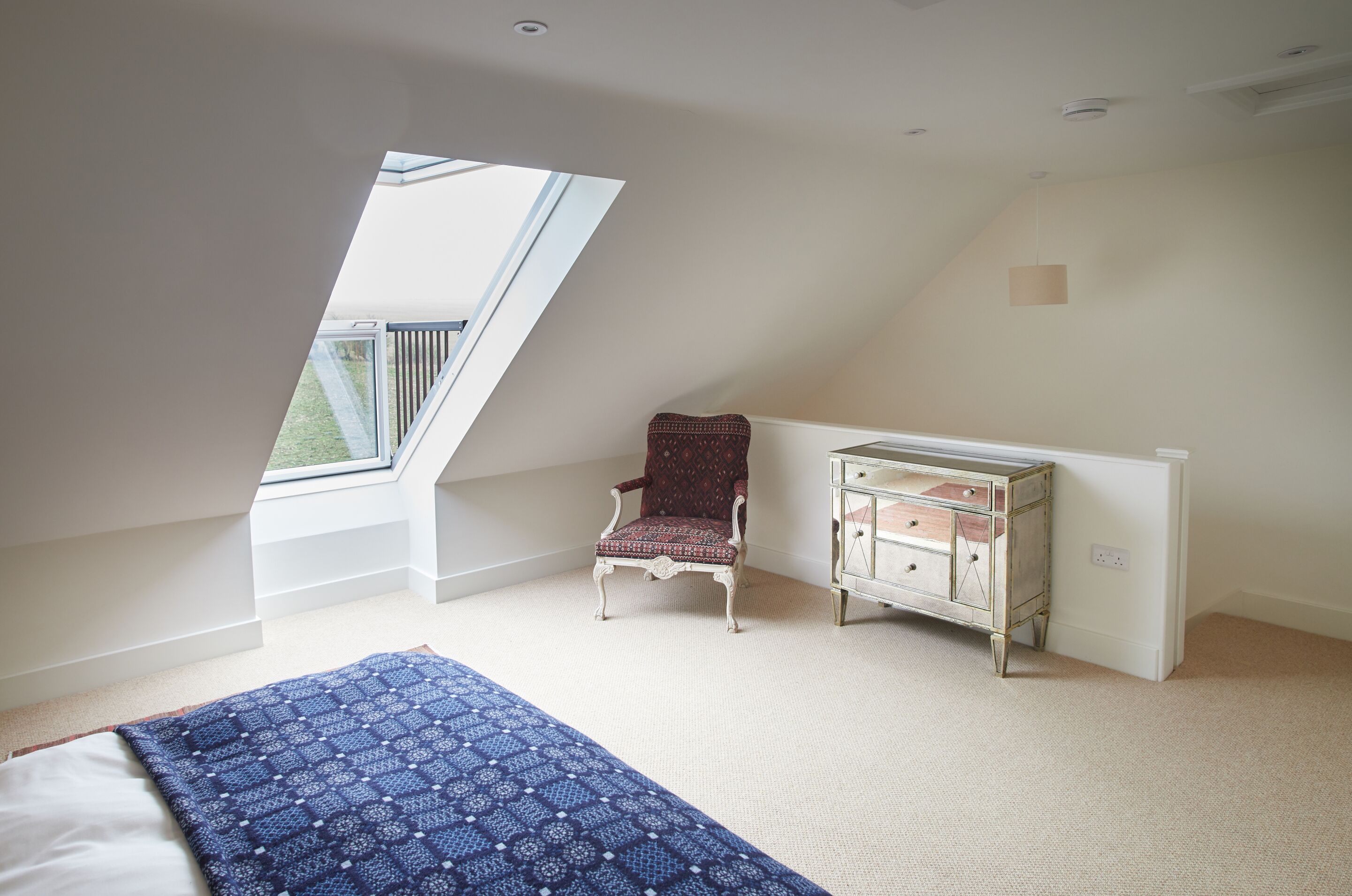 Une chambre avec un plafond en pente, une fenêtre, une couvre-lit à motifs, une chaise à motifs et une commode.