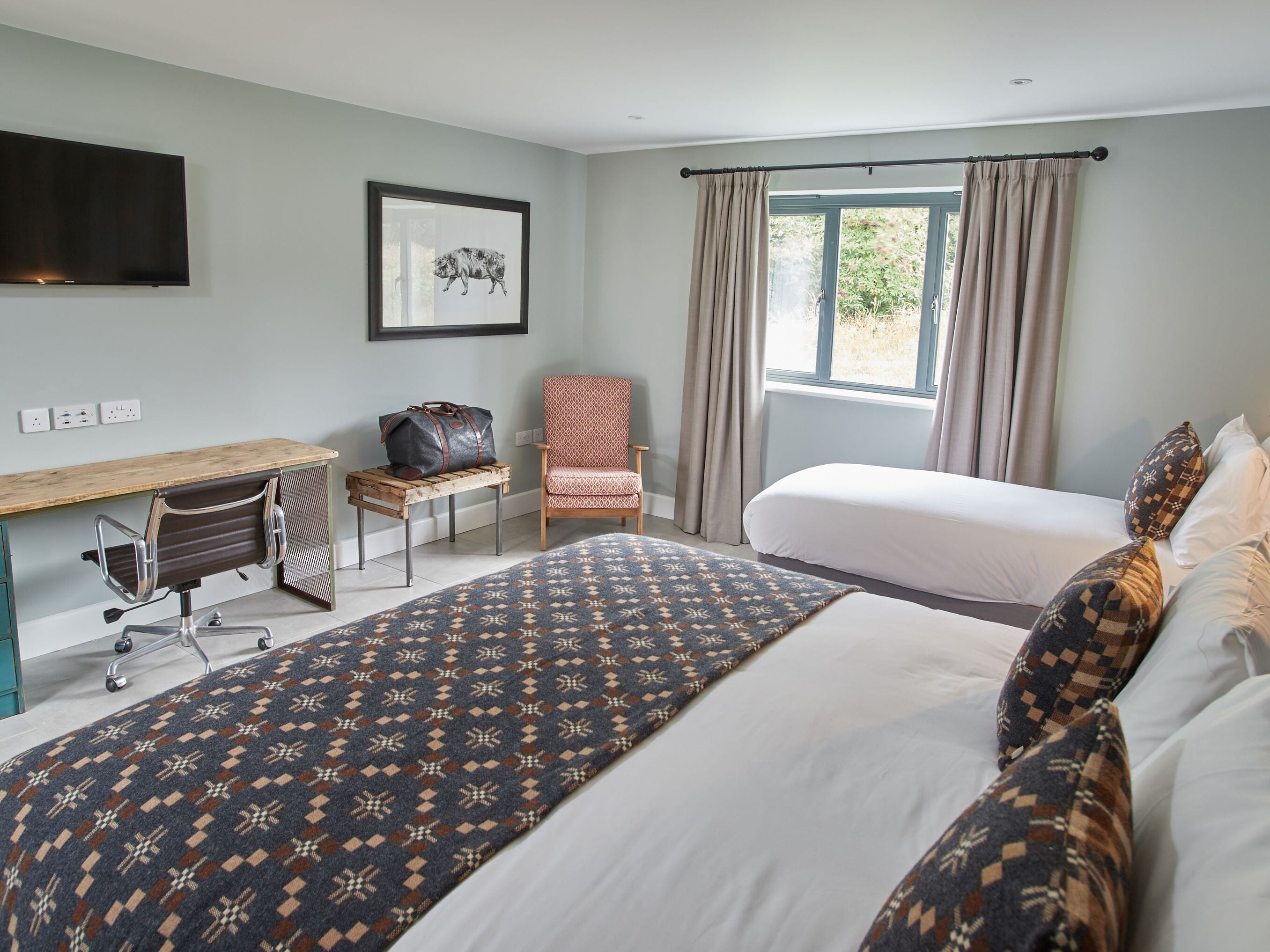 Habitación de hotel con dos camas, un escritorio con silla, un ventanal grande con cortinas y un televisor montado.