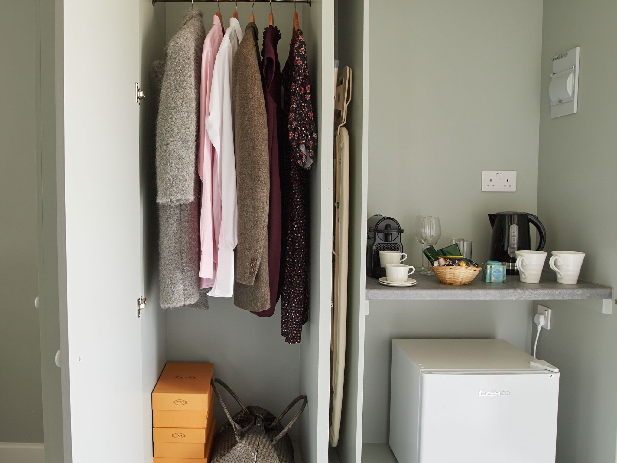 A compact kitchenette with a refrigerator, a small shelf with cups and a kettle, and a hanging clothes rack with various garments.