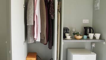 A compact kitchenette with a refrigerator, a small shelf with cups and a kettle, and a hanging clothes rack with various garments.