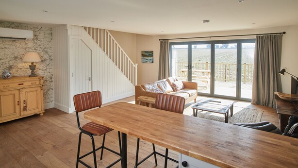 A spacious living area with a wooden dining table, a sofa, and a staircase leading to a balcony. - The Pig Shed Motel (King's Lynn)