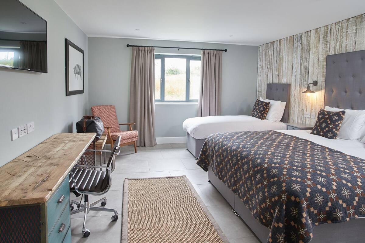 A hotel room with a bed, a desk, a chair, a window with curtains, and a headboard with a lamp.