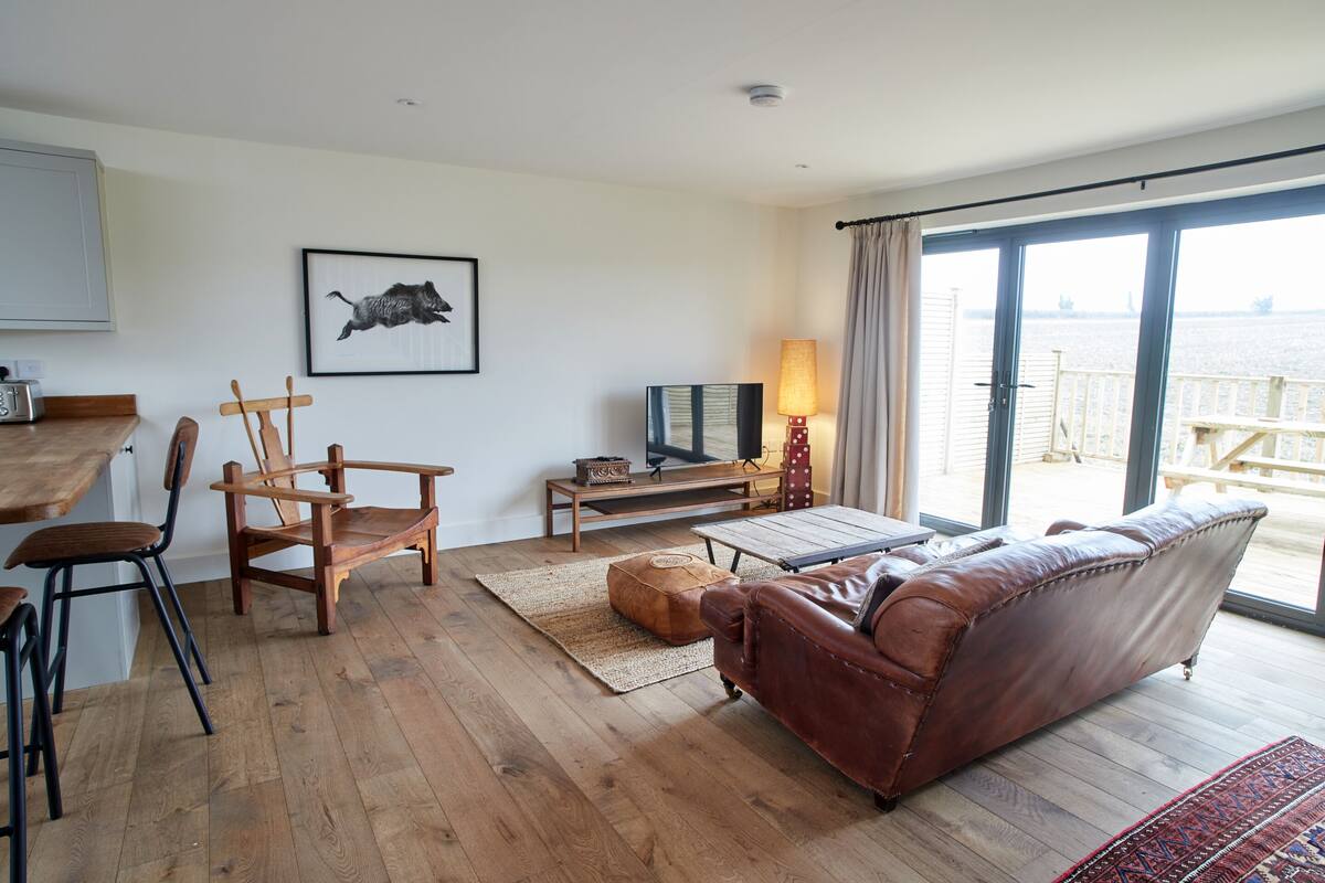 A living room with a wooden floor, a brown leather sofa, a wooden dining table with chairs, a TV, and a view of the sea through large windows.