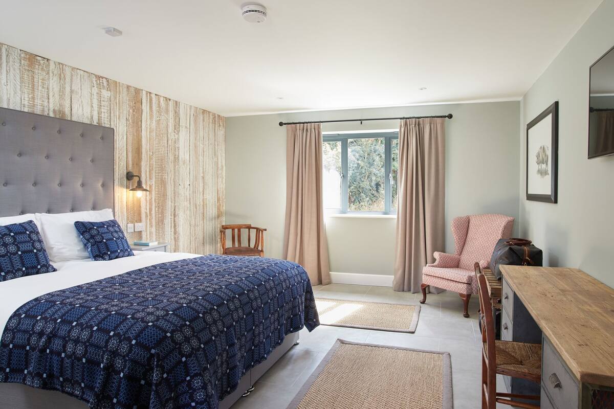 A bedroom with a large bed, a wooden desk, a chair, and a window with curtains.