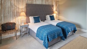 A hotel room with two beds, a wooden wall, a grey headboard, and a small bedside table with a lamp and books. - The Pig Shed Motel (King's Lynn)