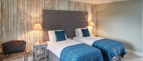 A hotel room with two beds, a wooden wall, a grey headboard, and a small bedside table with a lamp and books.