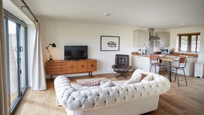 A living room with a sofa, a TV, a wooden cabinet, and a kitchen area with a counter and chairs. - The Pig Shed Motel (King's Lynn)