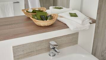 Un baño con tocador de madera, un lavabo blanco y un espejo. Hay dos cestas en el tocador, una con artículos de aseo y la otra con toallas dobladas.