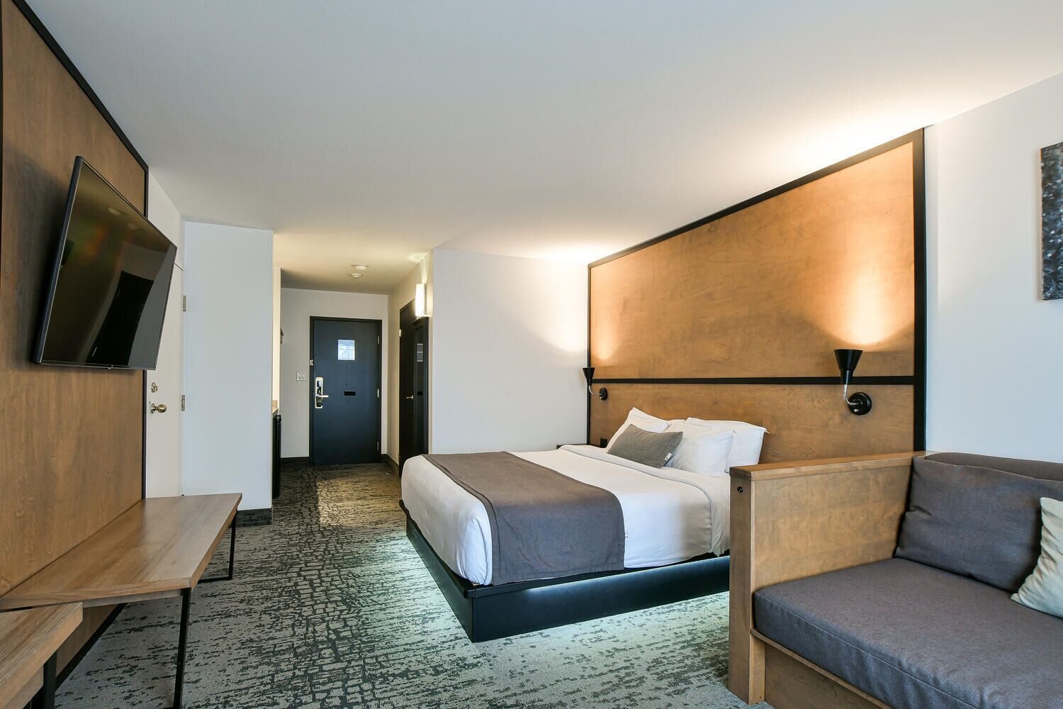 A modern hotel room with a bed, a seating area, a TV, and a hallway leading to a door.