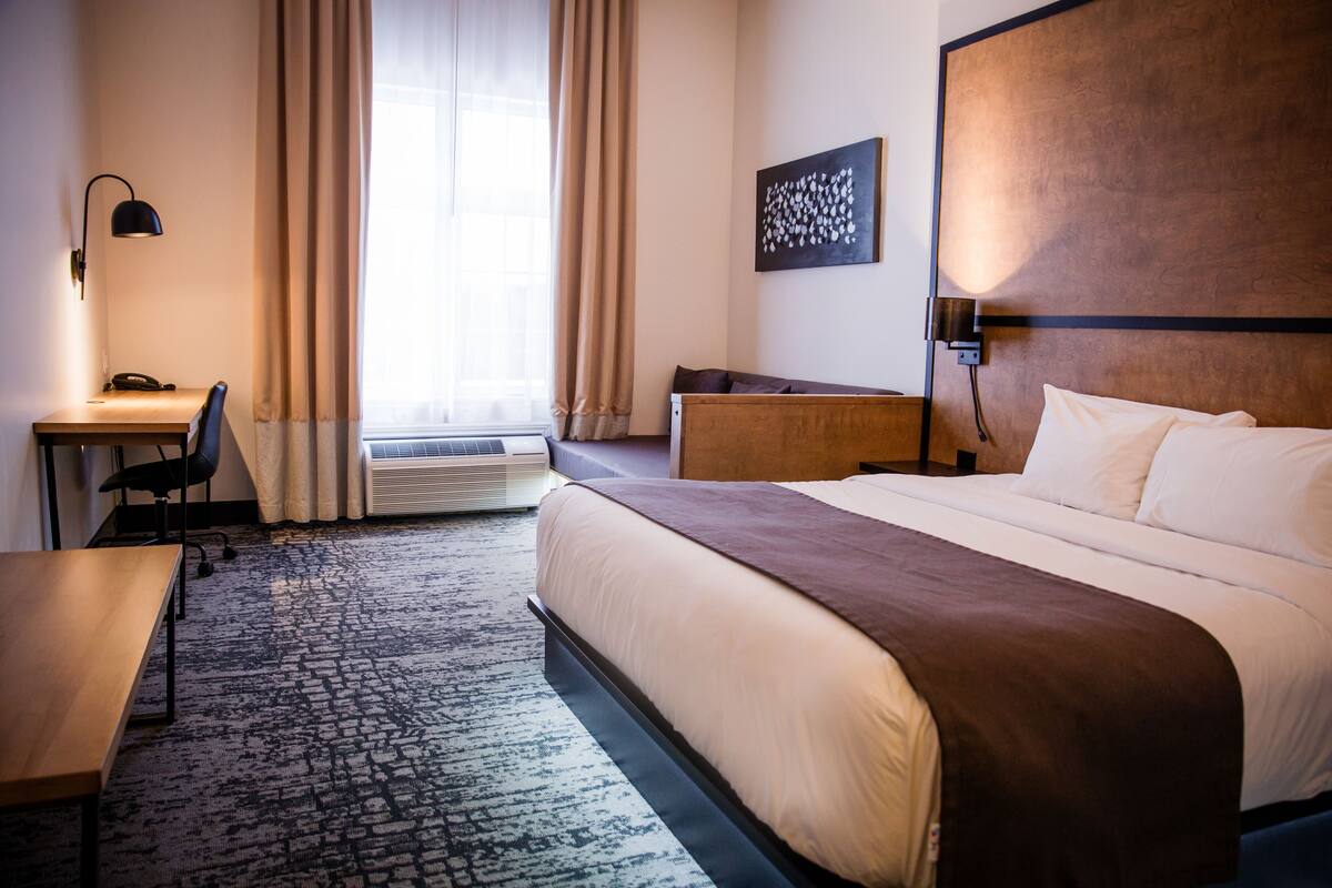 Una habitación de hotel con una cama grande, un escritorio, una silla, una lámpara y una ventana con cortinas.