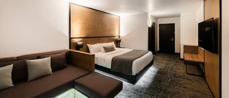 A modern hotel room with a bed, a sofa, a bedside table, a TV, and a carpeted floor.