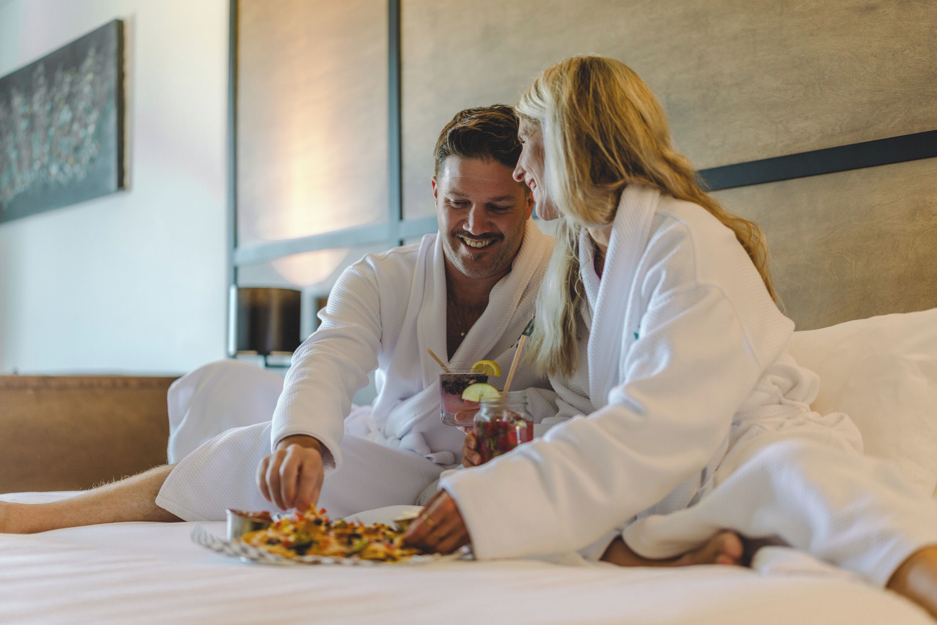 A couple in white bathrobes sitting on a bed with a plate of food and a drink.