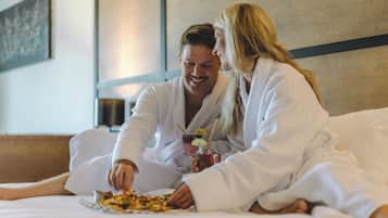 A couple in white bathrobes sitting on a bed with a plate of food and a drink.