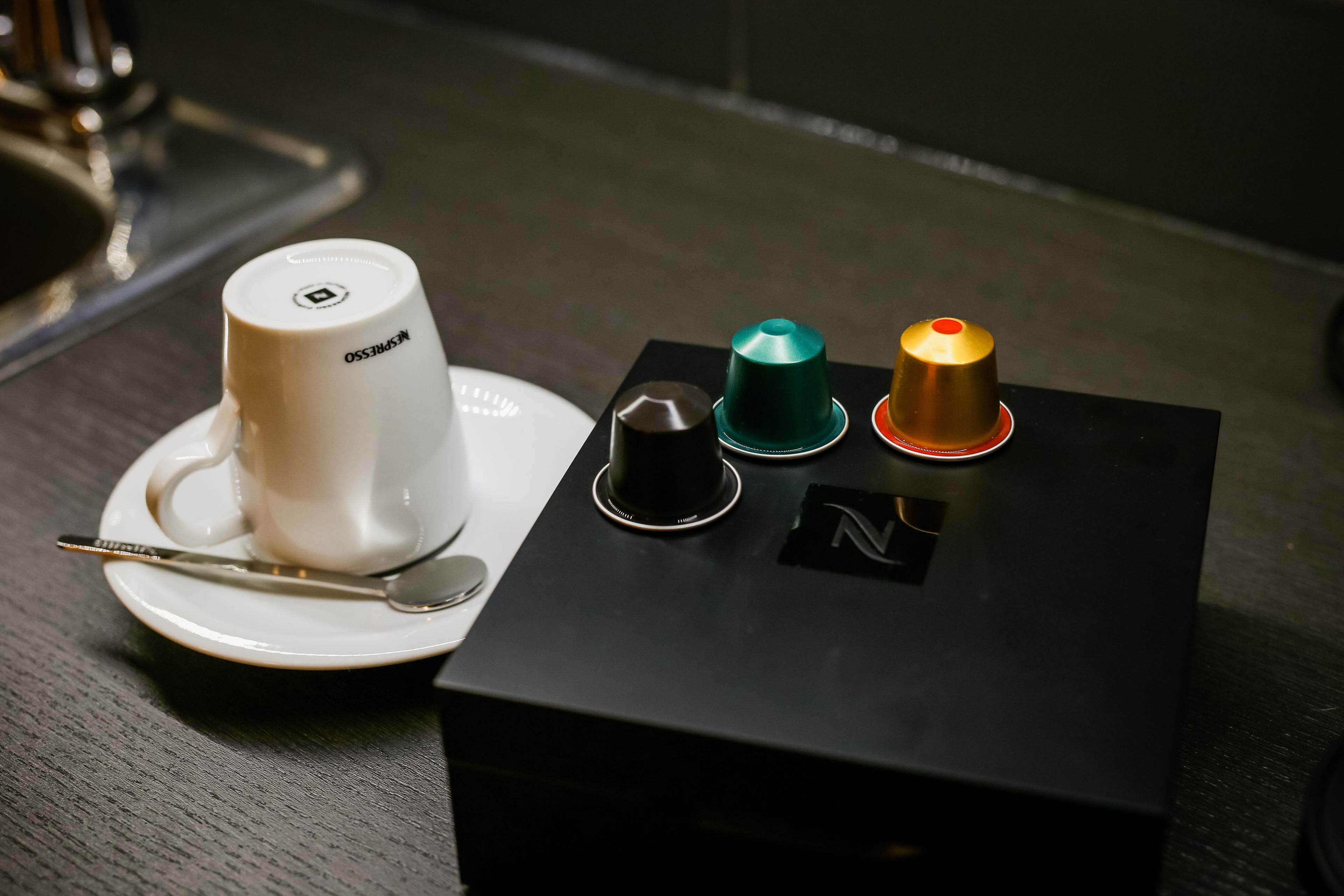 A coffee cup on a saucer with a spoon, a black box with three colored buttons, and a logo on the box.