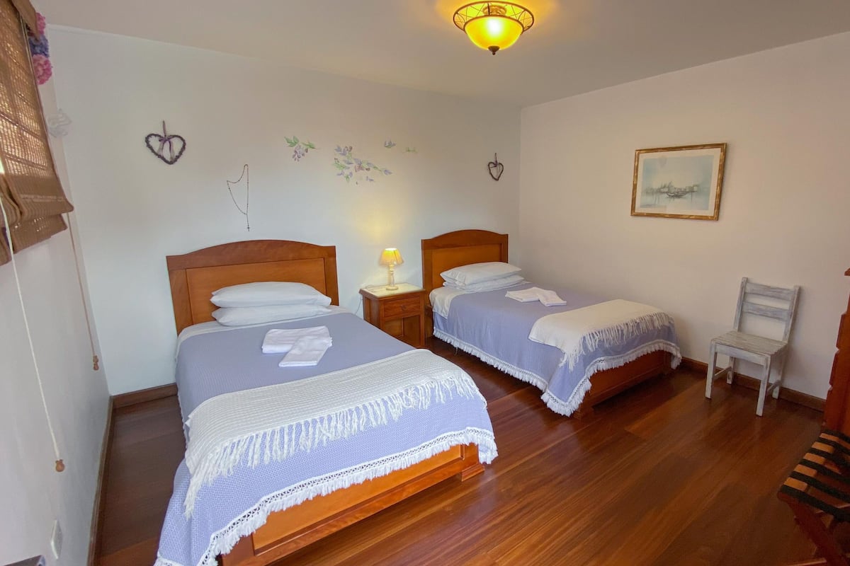 A hotel room with two beds, wooden flooring, a small table with a lamp, a chair, and a painting on the wall.