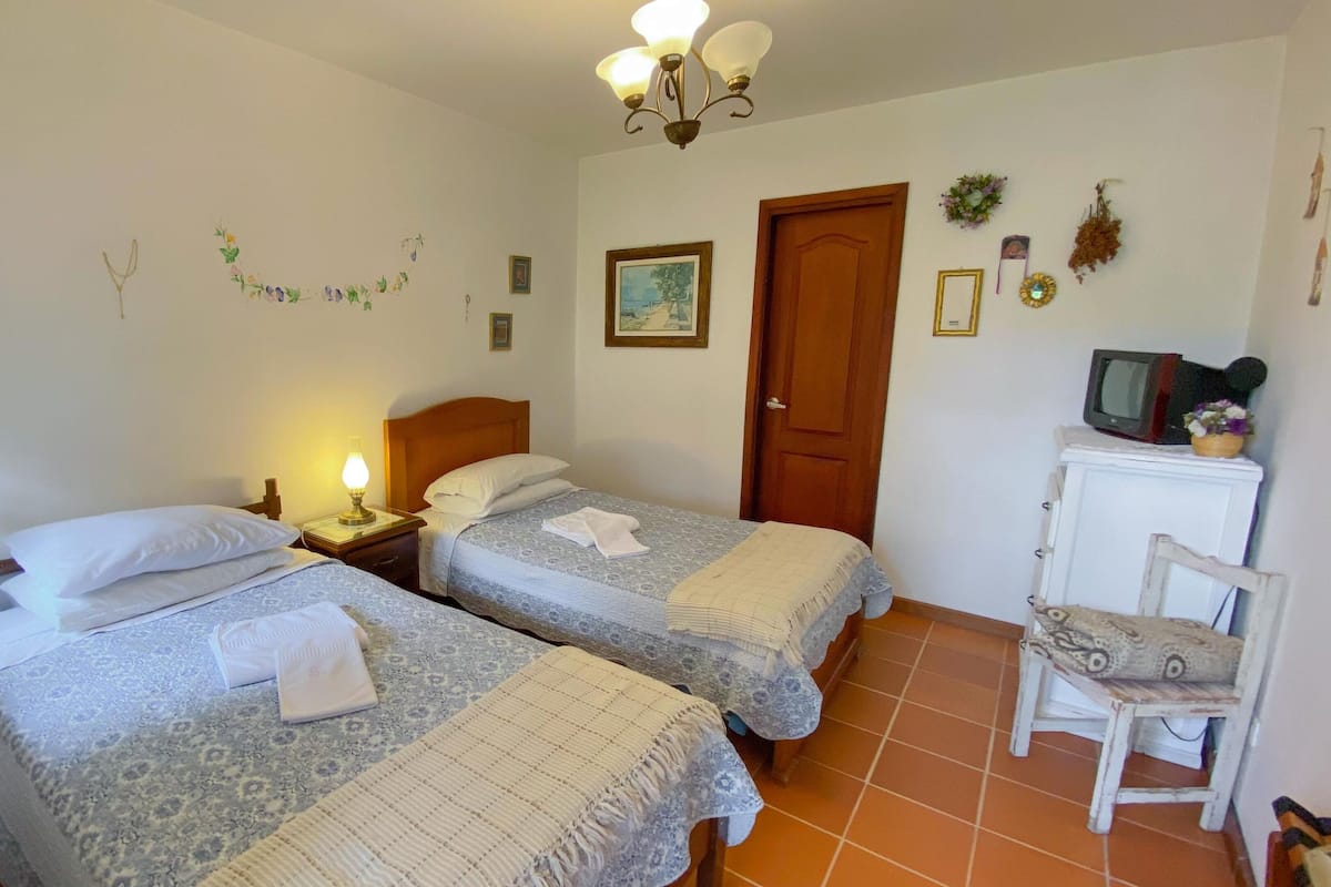 A hotel room with two beds, a nightstand with a lamp, a wooden door, a small white cabinet with a television, and a chair.