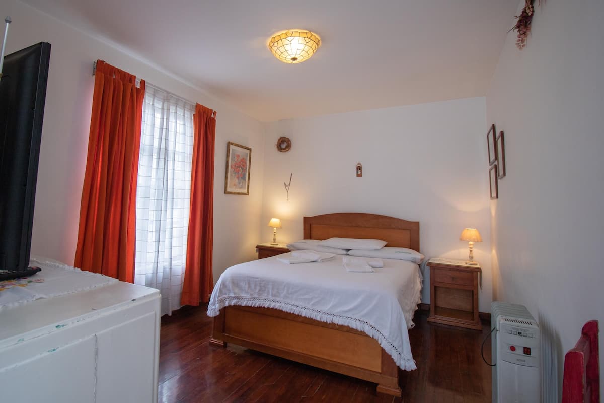 A bedroom with a wooden bed, two bedside tables with lamps, a heater, and a window with curtains.