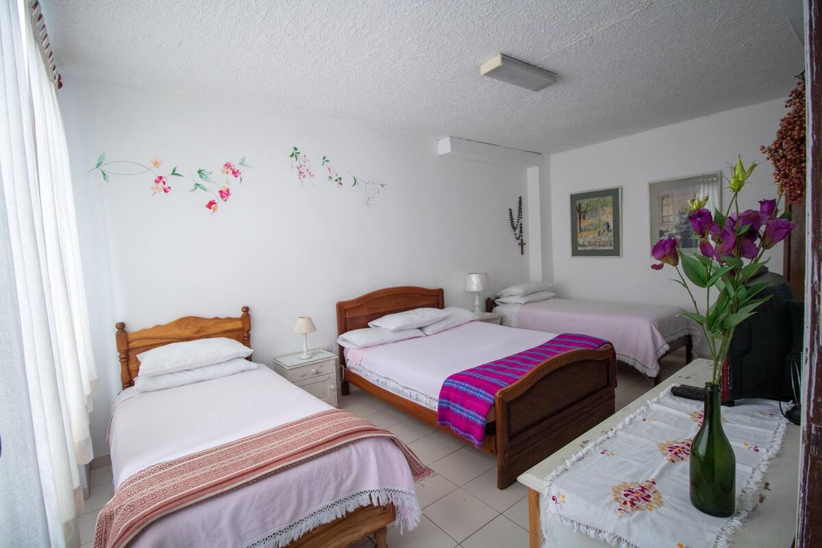A bedroom with two beds, a nightstand, a bedside table with a vase, and a wall with floral decorations.