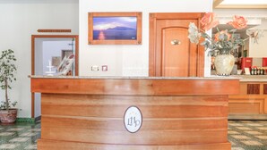A reception desk with a wooden finish, a framed picture on the wall, and a potted plant to the left. - Hotel La Pergoletta (Massa Lubrense)