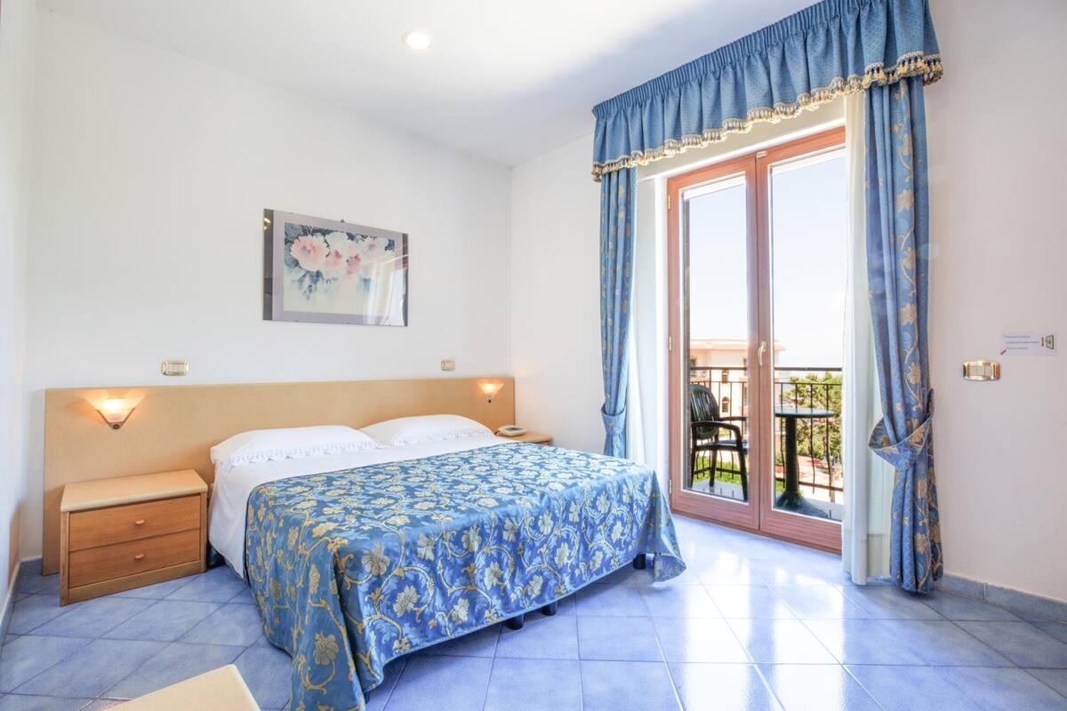A bedroom with a bed, bedside tables, a wall-mounted light, a window with curtains, and a balcony with a chair.