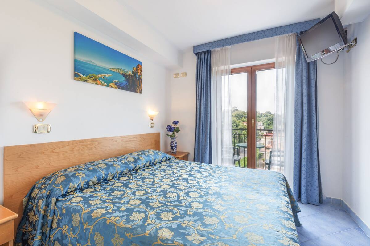 A bedroom with a large bed, a painting on the wall, a TV mounted on the wall, and a balcony with a table and chairs.