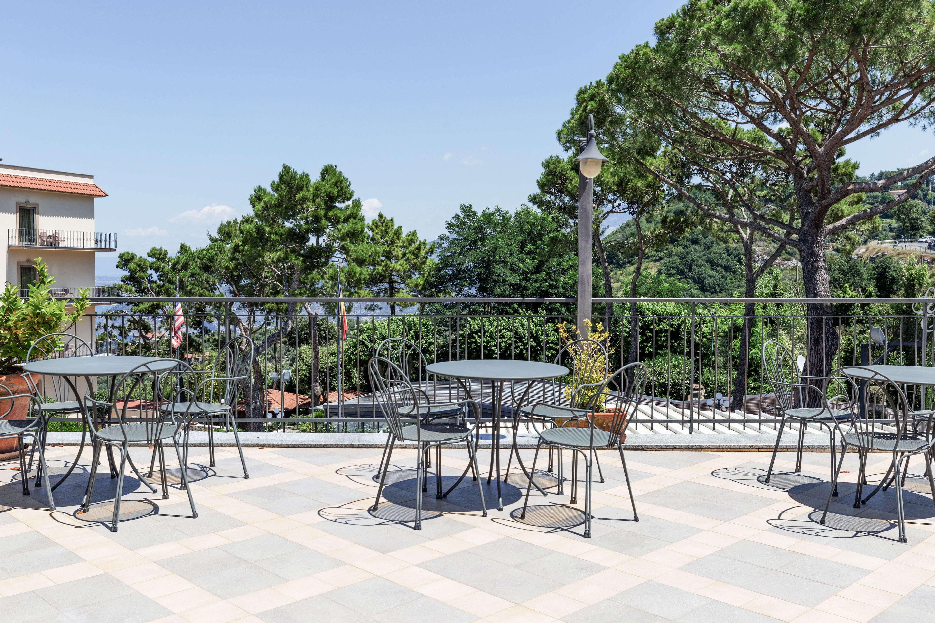 Eine Terrasse im Freien mit runden Tischen und Metallstühlen, umgeben von einem Metallzaun und Bäumen.