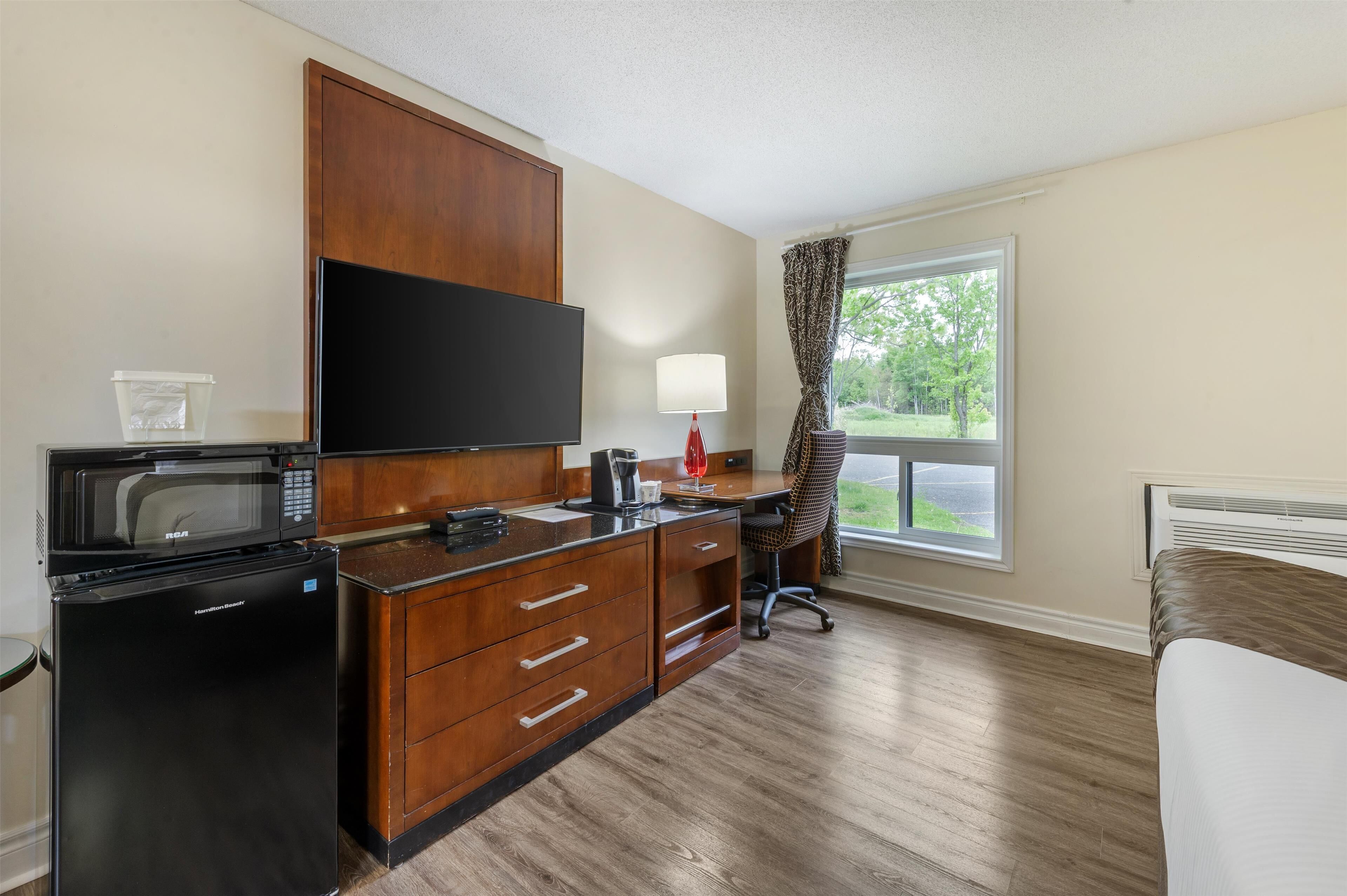 A compact hotel room with a kitchenette, a flat-screen TV, a desk with a chair, and a window with curtains.