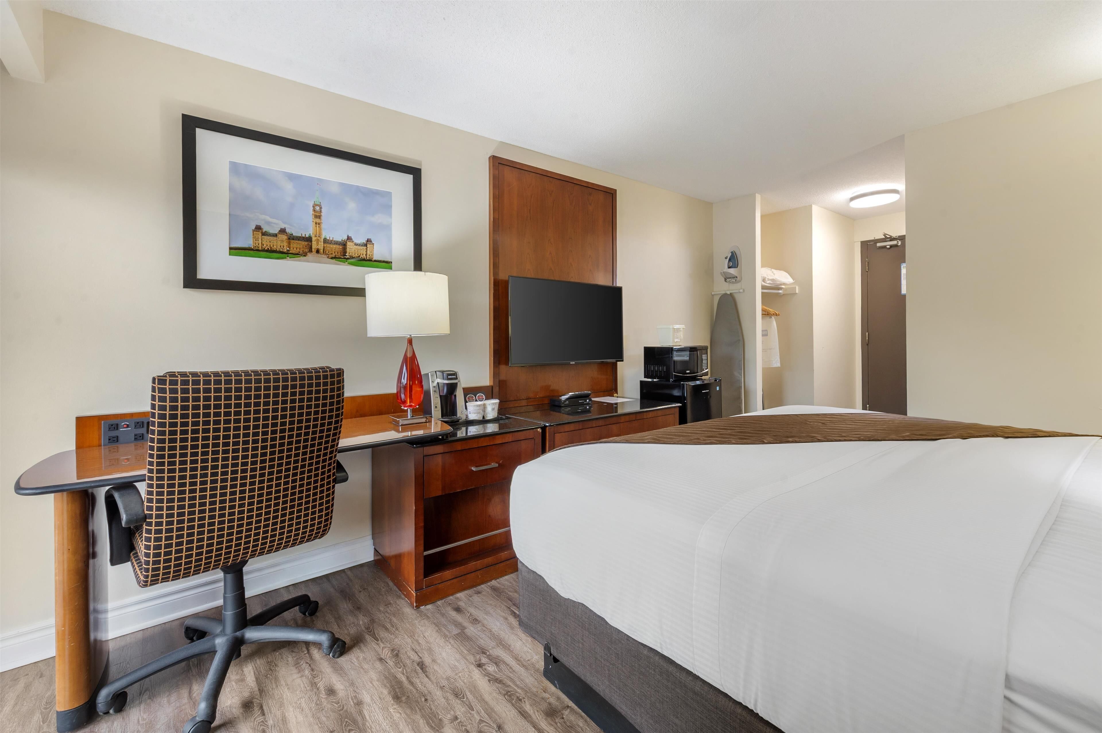 Une chambre d’hôtel comprenant un lit, un bureau avec une chaise, une télévision, une machine à café et un tableau encadré au mur.