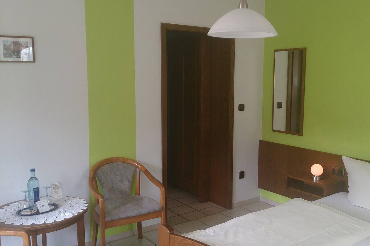 Une chambre d’hôtel avec un lit, une chaise, une petite table avec une bouteille et des verres, une table de chevet avec une lampe, et une porte donnant sur une autre pièce.