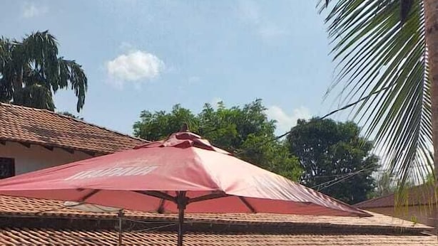Uma área de pátio com um guarda-sol vermelho, cadeiras de madeira e uma pequena mesa.