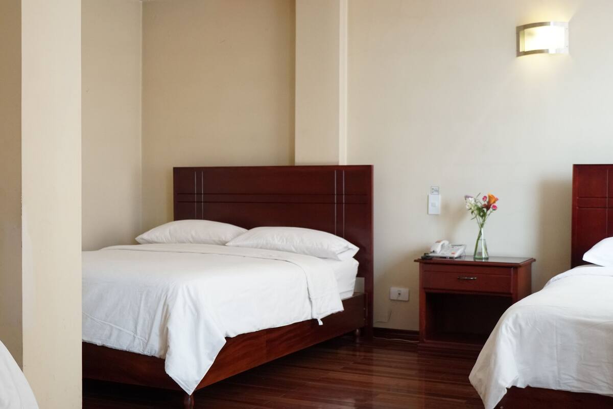 A hotel room with a wooden bed, a bedside table with a phone and vase, and a wall-mounted light.