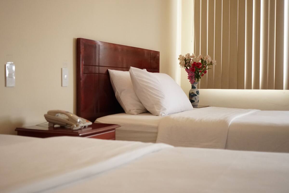 A hotel room with two beds, a wooden headboard, a bedside table with a telephone, and a vase with flowers.