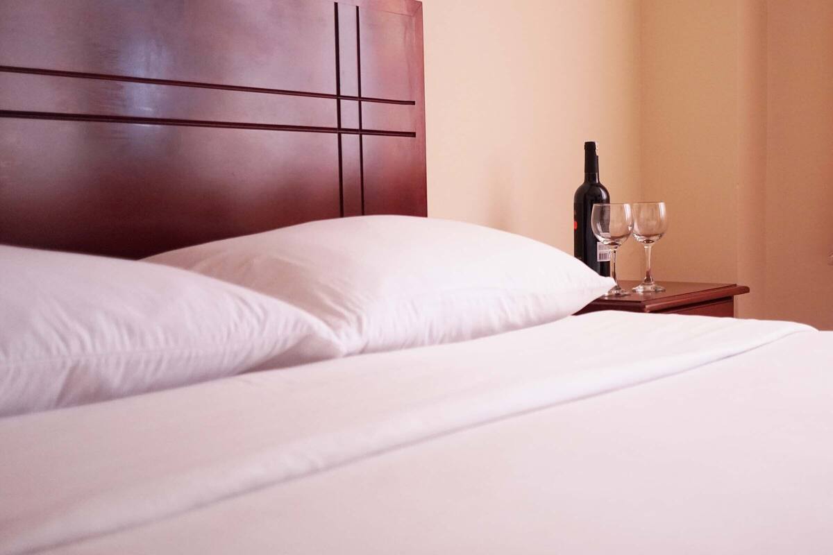 A neatly made bed with white linens and a wooden headboard. There is a small wooden nightstand beside the bed with a bottle and two glasses on it.