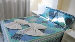 Una cama bien tendida con un edredón azul y verde, dos toallas dobladas encima y una mesita de noche blanca.