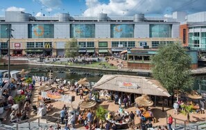 A bustling outdoor area with a river, seating areas, and a large building with commercial establishments.