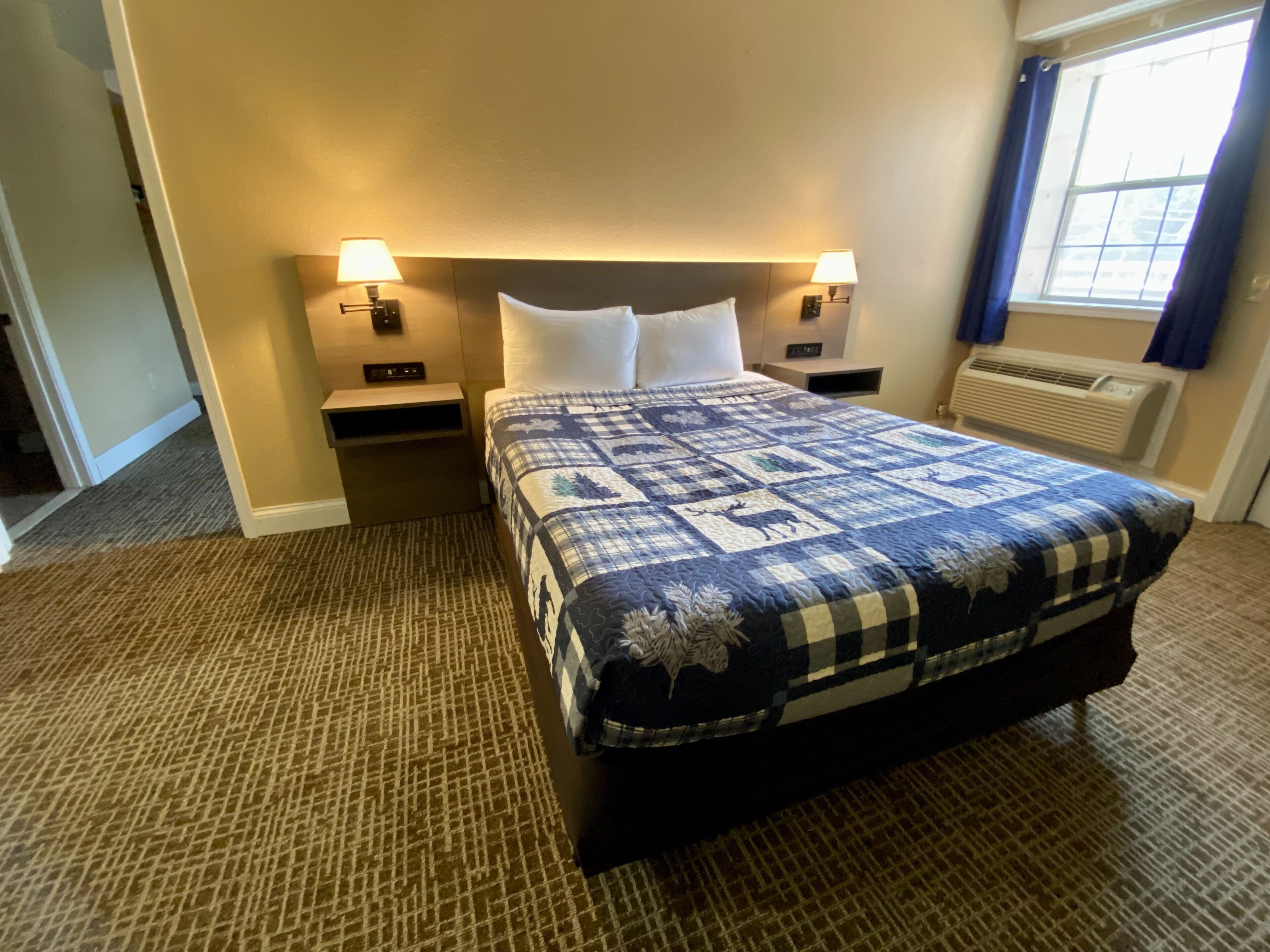 A hotel room with a large bed, two bedside tables, a nightstand, a window with curtains, and an air conditioning unit.