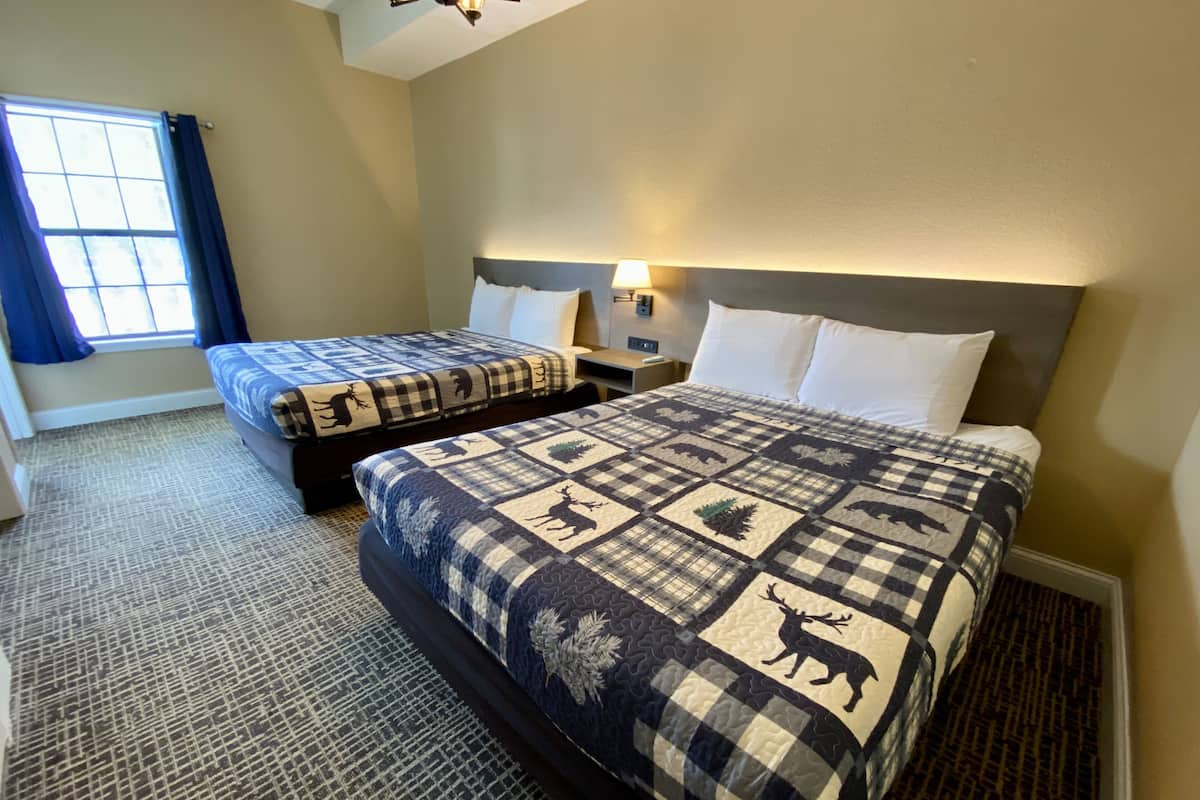 Habitación de hotel con dos camas, una ventana con cortinas, una mesita de noche y una lámpara en la pared.