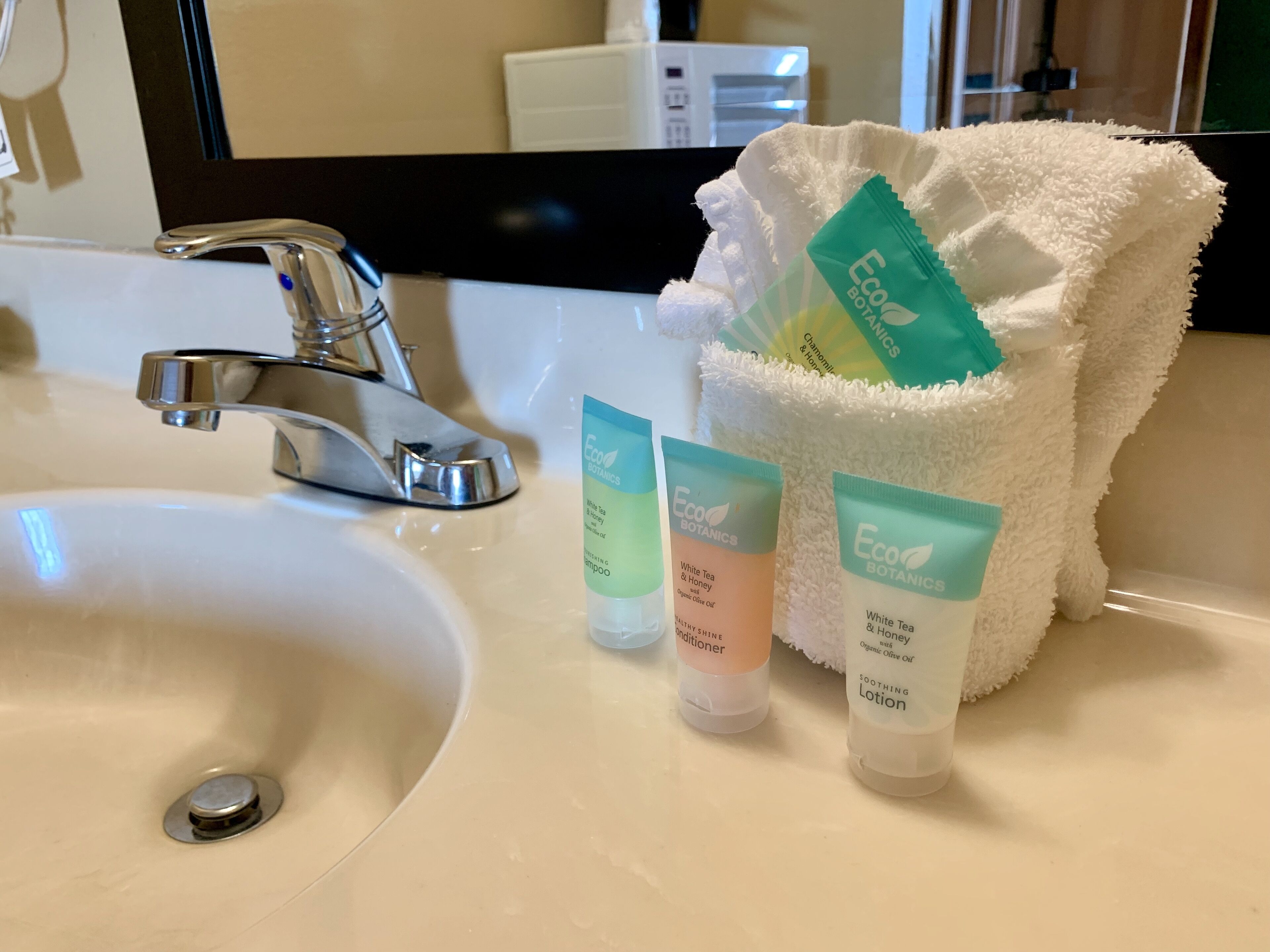 A bathroom sink with a chrome faucet, a white towel, and three Eco Botanics products: a white tea and honey conditioner, a white tea and honey soothing lotion, and a white tea and honey shampoo.