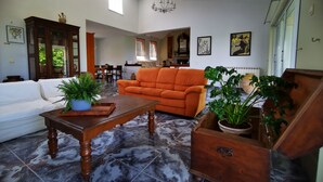 A spacious living room with a white sofa, an orange armchair, and a wooden coffee table. There are potted plants, a chandelier, and artwork on the walls. - Villa Irene (Kos)