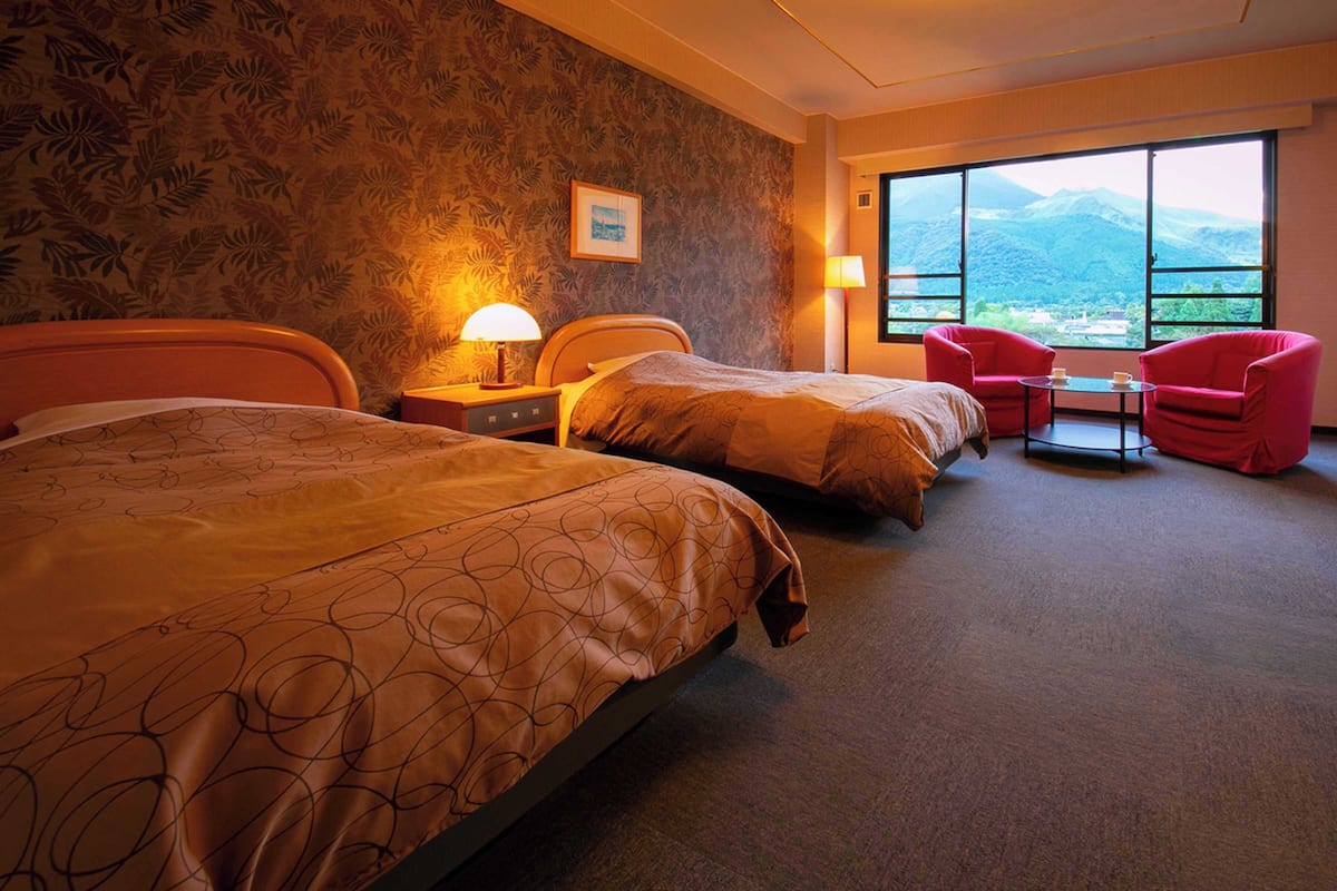 A hotel room with two beds, a small table, two chairs, and a large window offering a mountain view.