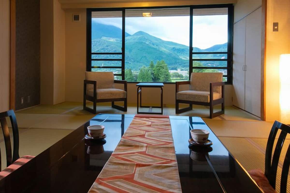 A traditional Japanese-style room with tatami flooring, a low table with a patterned runner, two chairs, and a view of mountains through large windows.