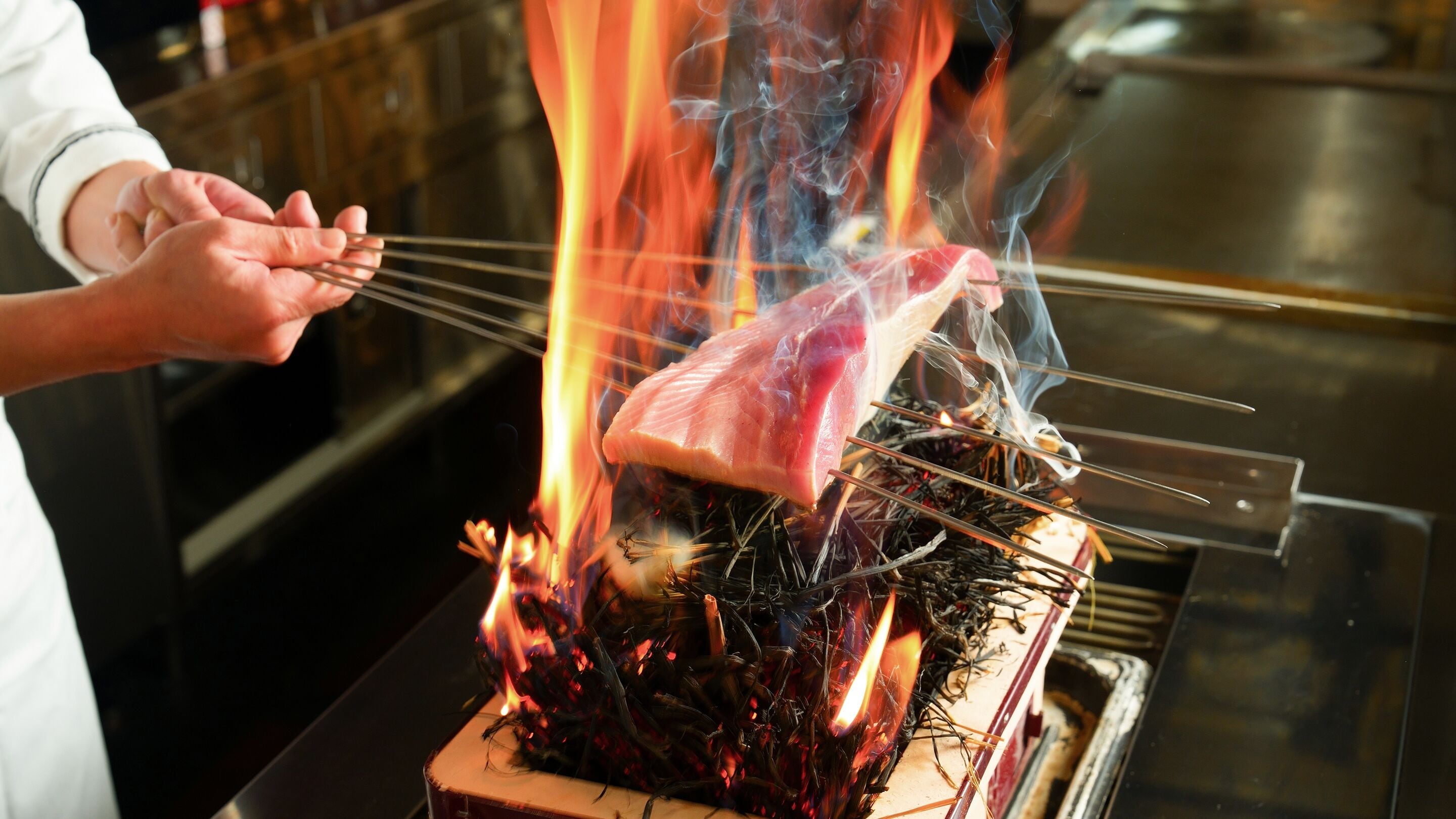 Un chef asando carne a la parrilla sobre fuego directo, usando brochetas.