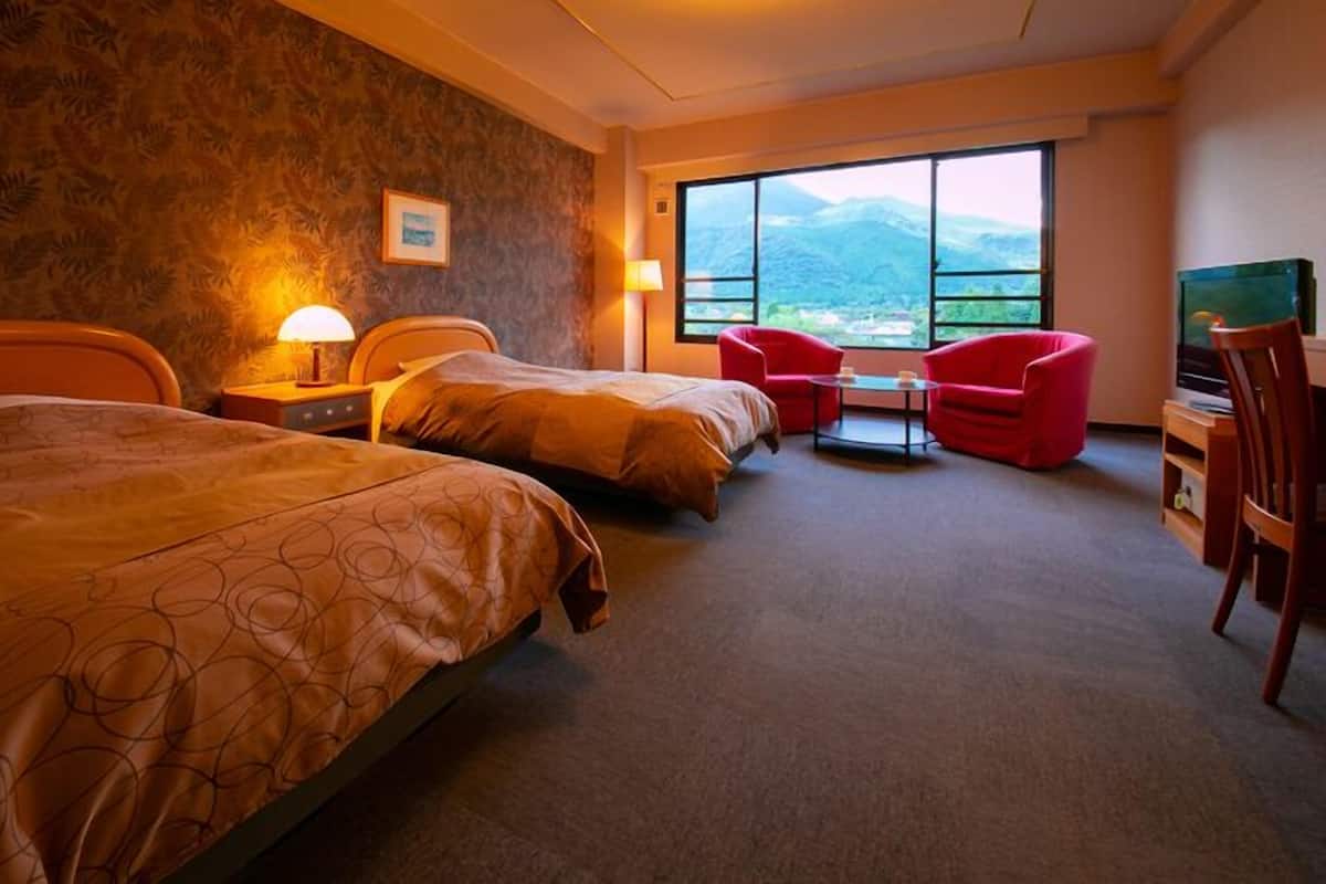 A hotel room with two beds, a desk with a chair, a TV, and a large window with a mountain view.