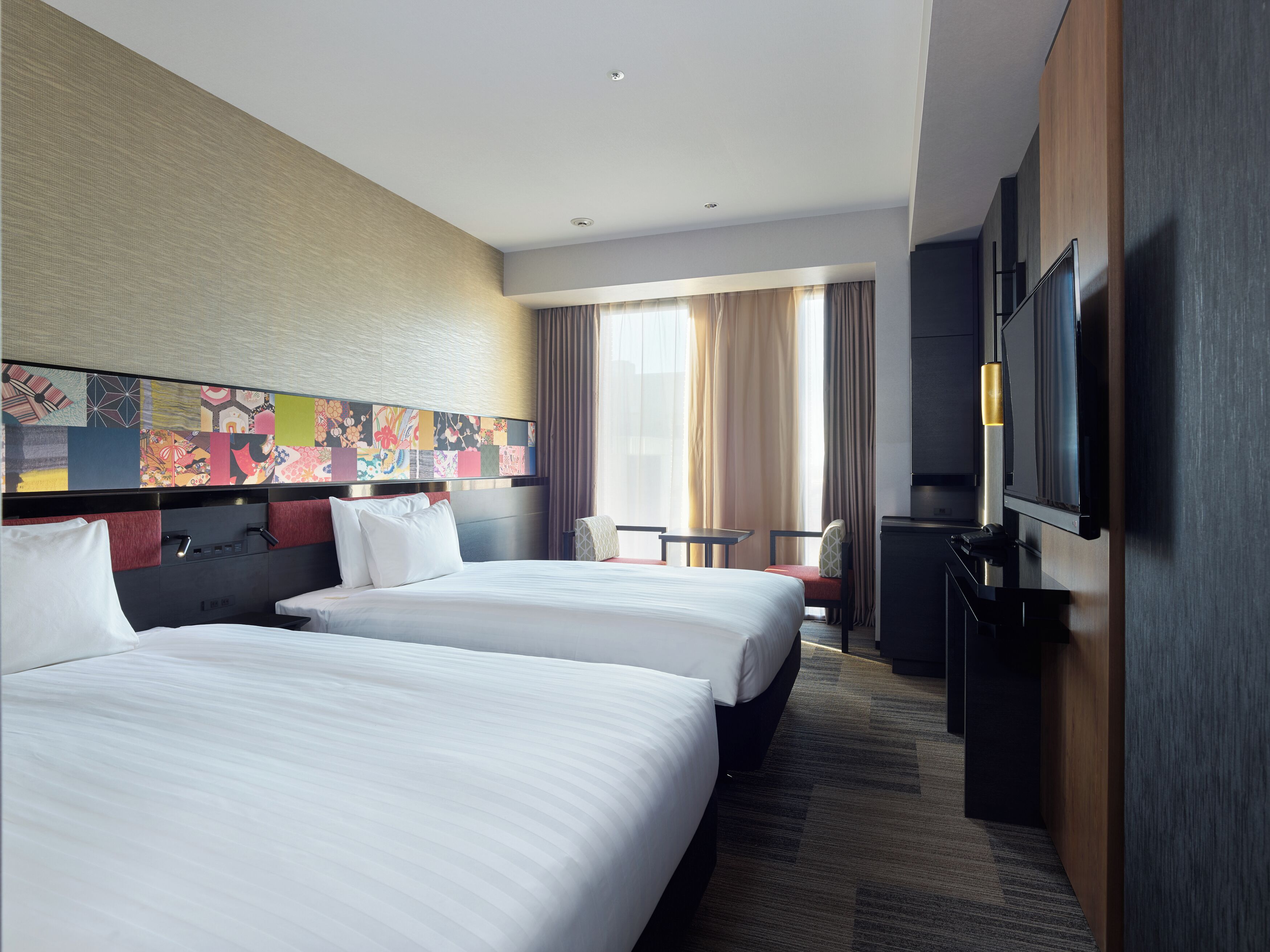 A hotel room with two beds, a TV, a desk, and a large window with curtains.